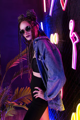 Beautiful girl with a cornrows hairstyle, wearing denim jacket and sunglasses, posing in a night club at the rabitz net wall, covered with neon lamps. Commercial, fashionable, conceptual design.