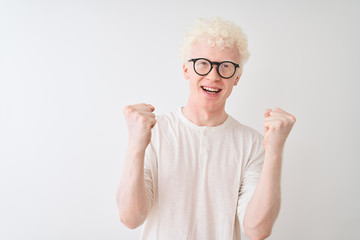 Young albino blond man wearing t-shirt and glasses standing over isolated white background celebrating surprised and amazed for success with arms raised and open eyes. Winner concept.