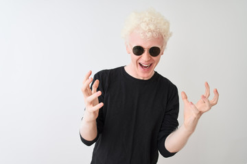 Young albino man wearing black t-shirt and sunglasess standing over isolated white background celebrating crazy and amazed for success with arms raised and open eyes screaming excited. Winner concept