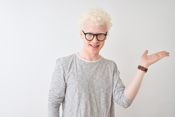Young albino blond man wearing striped t-shirt and glasses over isolated white background smiling cheerful presenting and pointing with palm of hand looking at the camera.