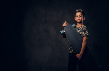 Small attractive boy is posing at dark photo studio with his skateboard on the dark background. © Fxquadro