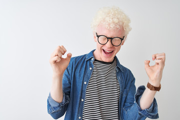 Young albino blond man wearing denim shirt and glasses over isolated white background celebrating mad and crazy for success with arms raised and closed eyes screaming excited. Winner concept