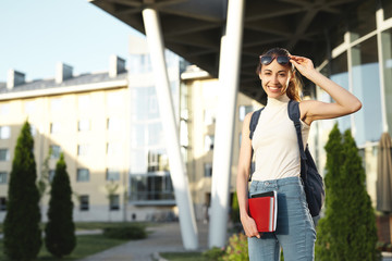 Beautiful happy woman in sunglasses with a backpack going to college. Young female university student with books in campus.