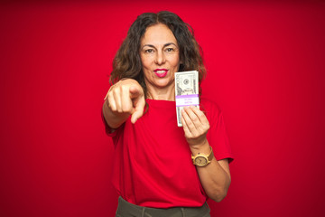 Middle age senior woman holding bunch of dollars over red isolated background pointing with finger to the camera and to you, hand sign, positive and confident gesture from the front