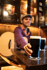 Man sitting on a table with a acoustic guitar taking a dark beer glass