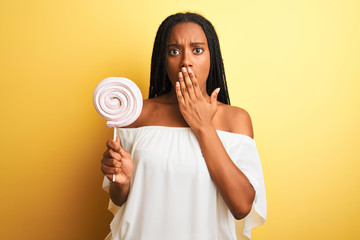 Young african american woman eating candy standing over isolated yellow background cover mouth with hand shocked with shame for mistake, expression of fear, scared in silence, secret concept