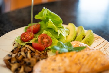 Close up of a grilled salmon fillet, a mushroom portion and a olive oil pouring on the salad.