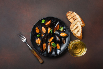 Seafood mussels and basil leaves in a black plate with wineglass, bread slices and fork on rusty background