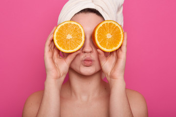 Horizontal shot of funny cheerful young woman holding two halves of orange against her eyes, having white towel on her head, posing with bared shoulders over rosy background. Beauty concept.