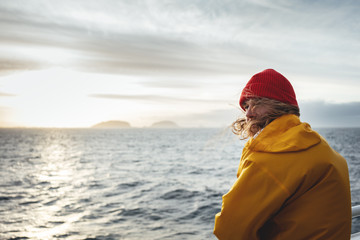 Hipster traveler wearing red hat and yellow raincoat looking away at cloudy mountain and sunset sea. Alone man traveling  at scandinavian authentic ocean landscape by ship