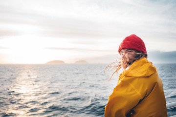 Fototapeta na wymiar Alone man traveling on ship and looking at sunset sea and foggy mountain on skyline. Hipster traveler wearing yellow raincoat and red hat enjoying beautiful ocean after storm