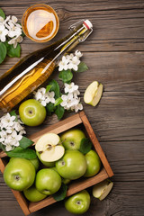 Ripe green apples in wooden box with branch of white flowers, glass and bottle of cider on a wooden table