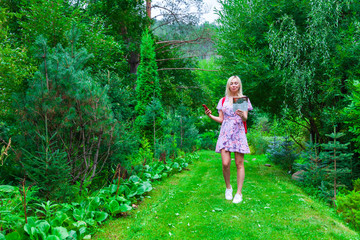 a young blonde girl in a pink dress with a red backpack on her back, a phone in her hand walks along a path and looking to the map in a park or forest. Travelling in wild.