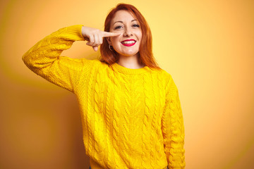 Beautiful redhead woman wearing winter sweater standing over isolated yellow background Pointing with hand finger to face and nose, smiling cheerful. Beauty concept