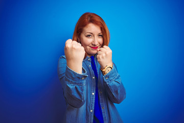 Young beautiful redhead woman wearing denim shirt standing over blue isolated background Ready to fight with fist defense gesture, angry and upset face, afraid of problem