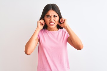 Young beautiful woman wearing pink casual t-shirt standing over isolated white background covering ears with fingers with annoyed expression for the noise of loud music. Deaf concept.