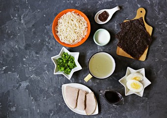Ingredients for making ramen soup on a dark concrete background: prepared ramen noodles, pork, miso paste, soy sauce, broth, chopped green onions, boiled egg, sugar, nori seaweed. Japanese food.