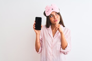 Young woman wearing sleep mask pajama holding smartphone over isolated white background serious...