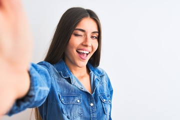 Beautiful woman wearing denim shirt make selfie by camera over isolated white background winking...