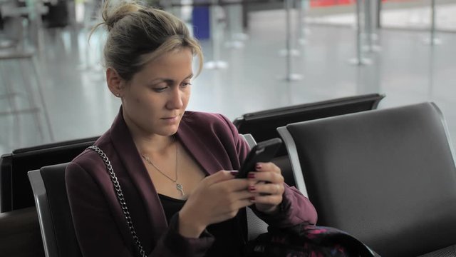 Attractive caucasian female typing message on smartphone while sitting at the airport terminal. Ulba and happy. The concept of waiting for the flight, vacation. Slow Motion, closeup.