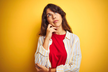 Young beautiful woman wearing red t-shirt and stripes shirt over yellow isolated background with hand on chin thinking about question, pensive expression. Smiling with thoughtful face. Doubt concept.