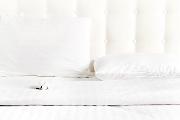 Soft white quilted pillow and cotton flower in bed on the background of white leather quilted headboard. Clean pillow, part of bed close-up, comfort. Quilted headboard background, bedding mockup