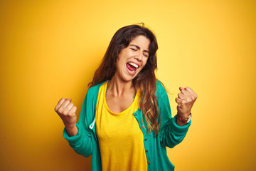 Young woman wearing t-shirt and green sweater standing over yelllow isolated background very happy and excited doing winner gesture with arms raised, smiling and screaming for success. 