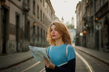 Young woman looking at the paper map and searching for direction early in the morning in ancient European city on empty street with tramway background