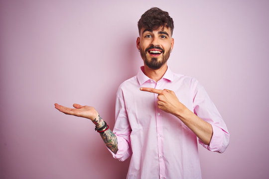 Young man with tattoo wearing shirt standing over isolated pink background amazed and smiling to the camera while presenting with hand and pointing with finger.