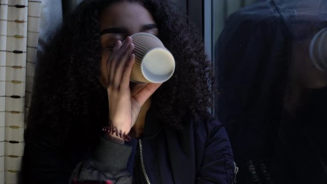 Sad depressed beautiful female mixed race African American girl teenager young woman teen wearing a black jacket sitting looking out of a window and drinking cup of coffee