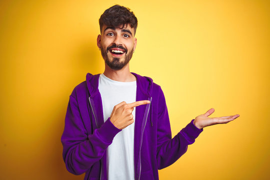 Young man with tattoo wearing sport purple sweatshirt over isolated yellow background amazed and smiling to the camera while presenting with hand and pointing with finger.