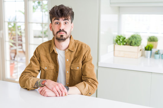 Young man wearing casual jacket sitting on white table puffing cheeks with funny face. Mouth inflated with air, crazy expression.