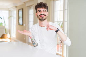 Young man wearing casual shirt sitting on white table amazed and smiling to the camera while presenting with hand and pointing with finger.