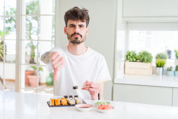 Young man eating asian sushi from home delivery with angry face, negative sign showing dislike with thumbs down, rejection concept