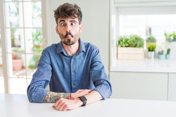 Young man wearing casual shirt sitting on white table making fish face with lips, crazy and comical gesture. Funny expression.