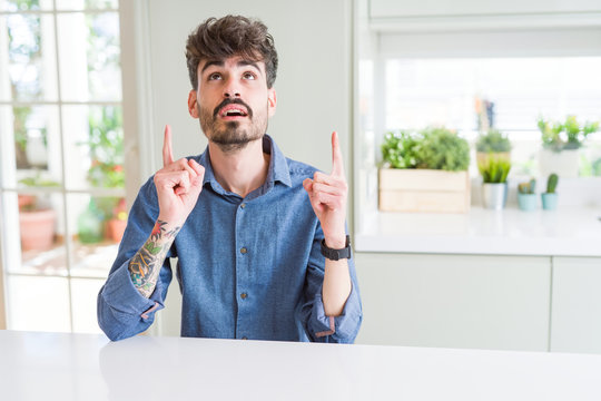Young man wearing casual shirt sitting on white table amazed and surprised looking up and pointing with fingers and raised arms.