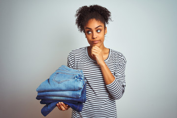 Young african american woman holding stack of jeans over isolated white background serious face thinking about question, very confused idea