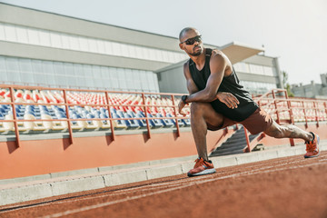 Fototapeta na wymiar Serious male athlete in sunglasses standing on the running track at the stadium while stretching his legs