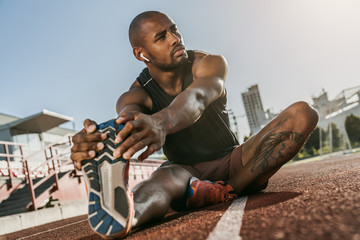 Sporty muscular african male athlete in earphones looking away, stretching legs while sitting at...