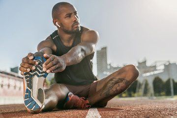 Sporty muscular african male athlete in earphones stretching legs while sitting at the stadium race...