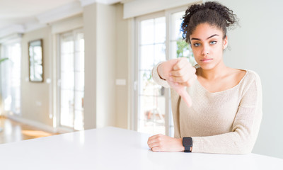 Beautiful young african american woman with afro hair sitting on table at home looking unhappy and angry showing rejection and negative with thumbs down gesture. Bad expression.