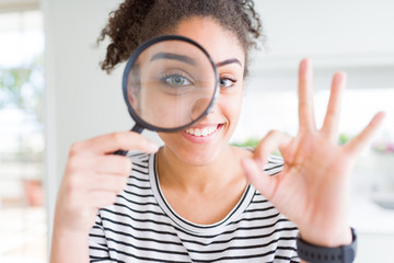 Young african american woman looking through magnifying glass doing ok sign with fingers, excellent...