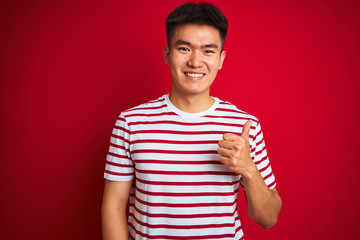 Young asian chinese man wearing striped t-shirt standing over isolated red background doing happy thumbs up gesture with hand. Approving expression looking at the camera showing success.