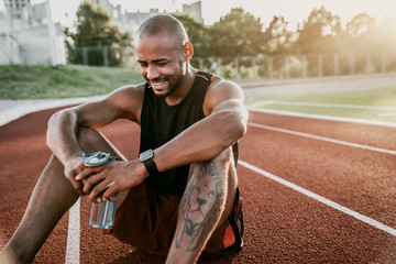 Cheerful young sporty man sitting on stadium running track with bottle of water in hands