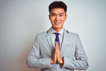 Asian chinese businessman wearing grey jacket and tie standing over isolated white background praying with hands together asking for forgiveness smiling confident.