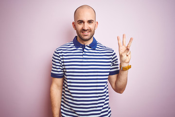 Young bald man with beard wearing casual striped blue t-shirt over pink isolated background showing and pointing up with fingers number three while smiling confident and happy.