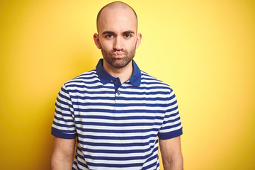 Fototapeta premium Young bald man with beard wearing casual striped blue t-shirt over yellow isolated background puffing cheeks with funny face. Mouth inflated with air, crazy expression.
