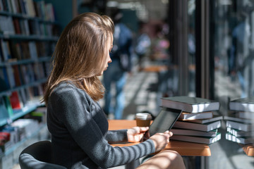 young caucasian teacher girl sitting in library at table with tablet pc and preparing for class