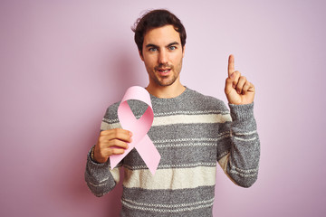 Young handsome man holding cancer ribbon standing over isolated pink background surprised with an idea or question pointing finger with happy face, number one