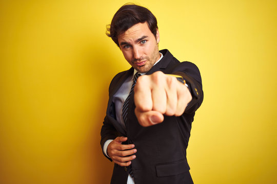 Young handsome businessman wearing suit and tie standing over isolated yellow background Punching fist to fight, aggressive and angry attack, threat and violence